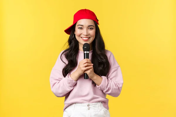İnsanların duyguları, yaşam tarzı, eğlence ve güzellik konsepti. Güzel gülümseyen Asyalı kadın şarkıcı kırmızı şapkalı şarkı söylüyor, mikrofonu elinde tutuyor rap yapıyor ya da şarkı söylüyor, sarı arka planda duruyor. — Stok fotoğraf