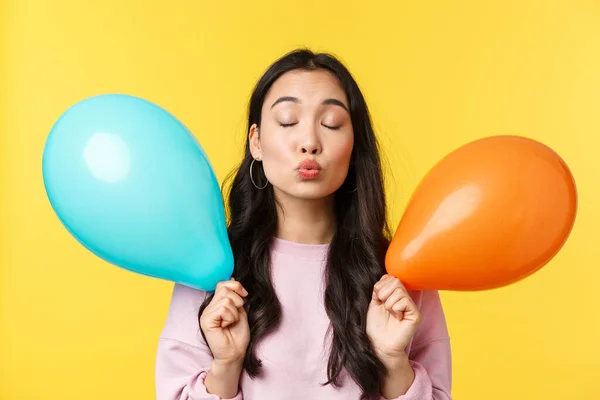 Persone emozioni, stile di vita concetto di svago. Sciocco e carino ragazza asiatica in possesso di due palloncini, in attesa di bacio con le labbra piegate e gli occhi chiusi, avendo momento romantico alla festa, sfondo giallo — Foto Stock