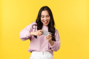 İnsanların duyguları, yaşam tarzı, eğlence ve güzellik konsepti. Neşeli hevesli Asyalı kız oyuncu cep telefonuyla oynuyor, akıllı telefonu oyun seviyesini geçmek için eğiyor, gülümseyerek eğleniyor.