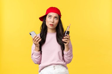 İnsanların duyguları, içkileri ve yaz tatili konsepti. Sinirli ve cahil Asyalı kadın, kırmızı şapkalı, ezikçe mesajlar okuyor, cep telefonu kullanıyor, gazoz içiyor, gazoz içiyor, sarı arka plan