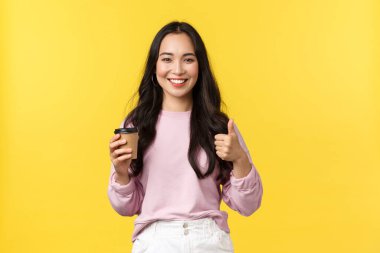 İnsanların duyguları, yaşam tarzı, eğlence ve güzellik konsepti. Gülümseyen Asyalı güzel kız, bardakları kaldırıyor, başparmaklarını kaldırıyor, nefis kahve ve tatlı ile harika bir kafe tavsiye ediyor, sarı arka planda.