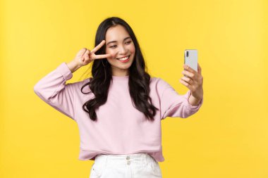 İnsanların duyguları, yaşam tarzı, eğlence ve güzellik konsepti. Sarı arka plandaki neşeli Asyalı kız cep telefonuyla selfie çekiyor, fotoğraf filtresi uygulaması kullanıyor ve yüzün yanında barış işareti ve gülümsüyor.
