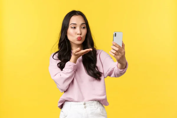 Persone emozioni, stile di vita tempo libero e concetto di bellezza. Elegante e flirty ragazza asiatica scattare foto per i social media, fare selfie, l'invio di baci aerei a fotocamera smartphone, stand sfondo giallo — Foto Stock