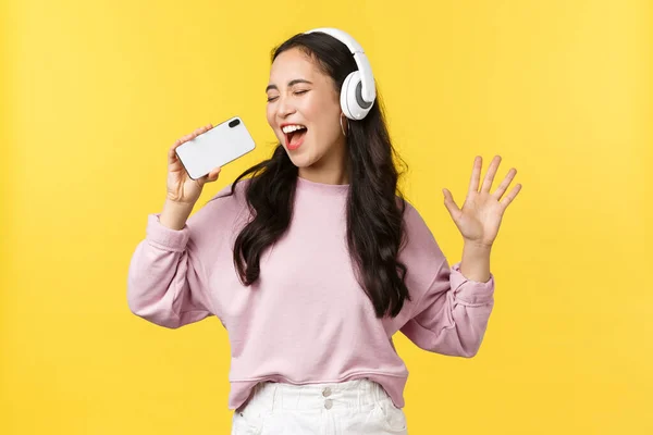 Gente emociones, estilo de vida ocio y concepto de belleza. Despreocupado mujer asiática feliz en los auriculares, jugando aplicación de karaoke en el teléfono móvil, utilizando el teléfono inteligente como micrófono y cantando a lo largo de la canción — Foto de Stock