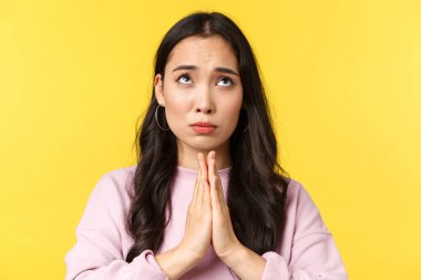 İnsanların duyguları, yaşam tarzı, eğlence ve güzellik konsepti. Umut dolu Asyalı kızın yakın çekimi, dua etmek için el ele tutuşması ve umutsuzca üzgün bir ifadeyle yukarı bakması, sarı arka plan