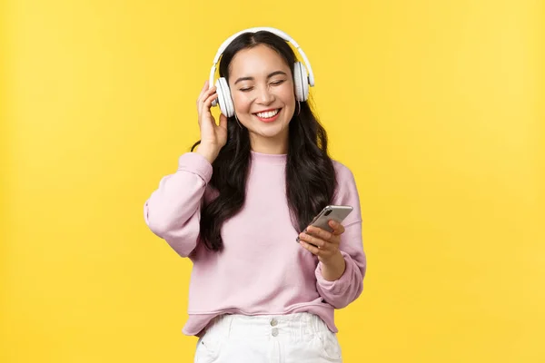 Gente emociones, estilo de vida ocio y concepto de belleza. Sonriendo despreocupada chica asiática moderna escuchando música en auriculares inalámbricos. Mujer disfrutando de la canción favorita en los auriculares, sosteniendo el teléfono móvil — Foto de Stock