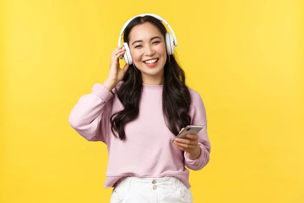 Gente emociones, estilo de vida ocio y concepto de belleza. Feliz sonriente mujer asiática escuchando música en auriculares inalámbricos y mirando complacido con buen sonido, sosteniendo el teléfono móvil — Foto de Stock