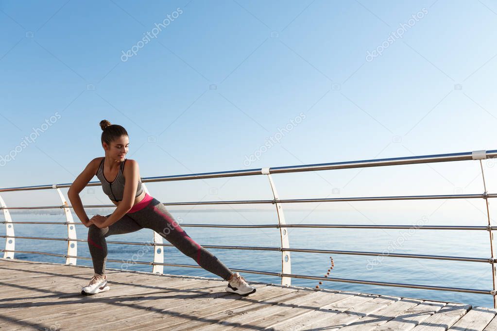 Young attractive sportswoman workout near the sea. Fitness woman stretching her legs before jogging and training on the seaside