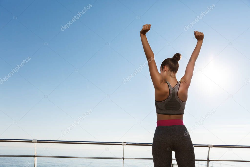 Rear view of happy triumphing sportswoman standing on a pier, raising hands up like champion, achieve goal during training session outdoors