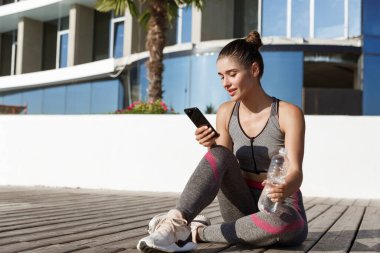 Açık havada, çekici fitness kadınının ahşap iskelede akıllı telefonuyla otururken, antremandan sonra mola verirken, su içerken.