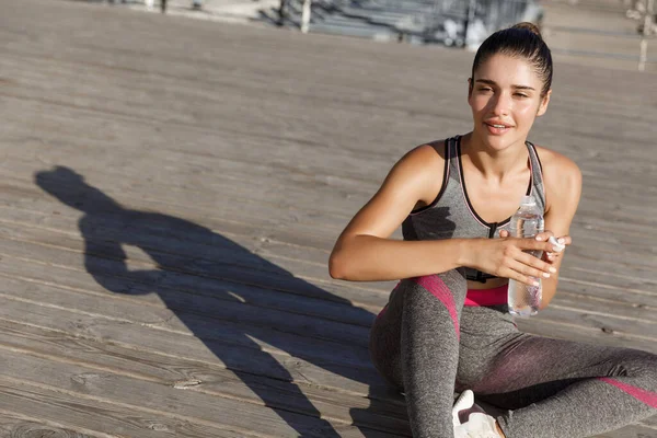 Primer plano de la atractiva deportista tomando un descanso después del entrenamiento, sentado a la orilla del mar y bebiendo agua, mirando complacido — Foto de Stock