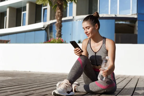 Foto al aire libre de atractiva mujer fitness sentada en el muelle de madera con teléfono inteligente, teniendo descanso después del entrenamiento, agua potable — Foto de Stock