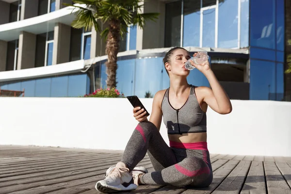 Foto al aire libre de atractiva mujer fitness sentada en el muelle de madera con teléfono inteligente, teniendo descanso después del entrenamiento, agua potable — Foto de Stock