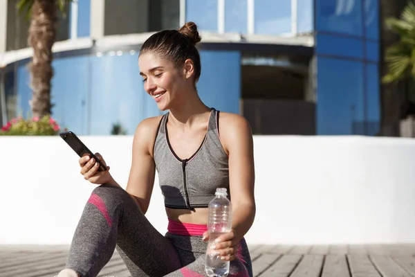 Foto al aire libre de la mujer de fitness feliz mirando la aplicación del teléfono móvil, sentado en el suelo de madera y agua potable — Foto de Stock