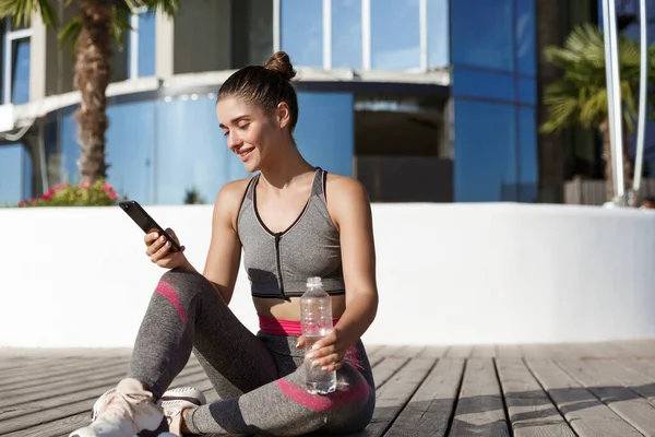 Foto al aire libre de la deportista atractiva feliz sentada en el suelo después del entrenamiento, agua potable y mirando el teléfono inteligente — Foto de Stock