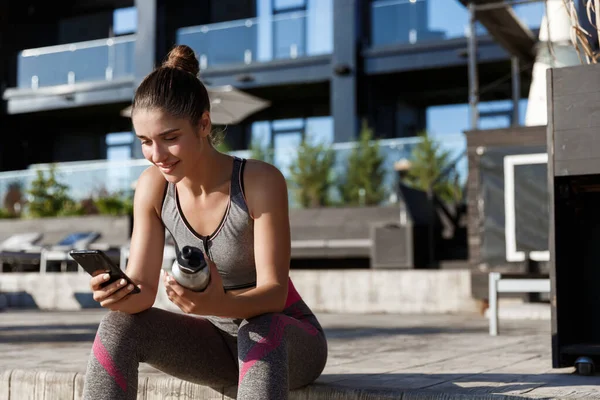 Foto al aire libre de la atractiva deportista sentada en el banco y usando el teléfono móvil, sosteniendo la botella de agua — Foto de Stock