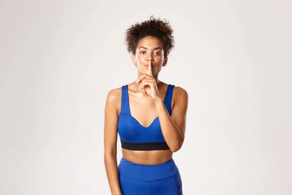 Jong Afrikaans-amerikaans sportief meisje in blauwe sport outfit, vragen stem laag, stil op camera, staande tegen een witte achtergrond — Stockfoto