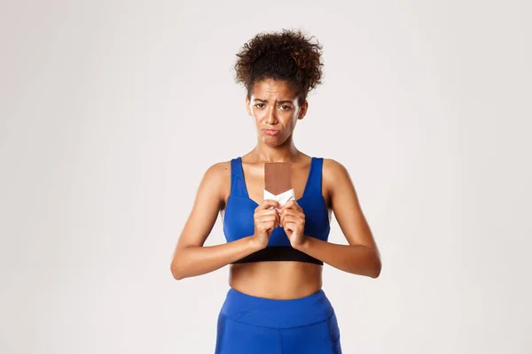 Droevig en somber Afrikaans-Amerikaans fitnessmeisje op dieet, met chocoladereep en grimmige overstuur, staande tegen een witte achtergrond — Stockfoto