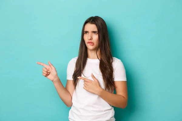 Immagine di bella donna perplessa in t-shirt bianca, cipiglio e labbro mordente complicato, indicando e guardando a sinistra lo spazio copia, in piedi su sfondo blu — Foto Stock