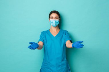 Covid-19, salgın hastalık ve ilaç konsepti. Tıbbi maskeli, eldivenli ve önlüklü güzel bayan doktor, hastayla ilgilenmek için ellerini uzatıyor, mavi arka planda duruyor.