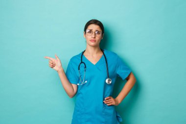 Coronavirus, salgın hastalık ve sosyalleşme konsepti. Gözlüklü, önlüklü, solcu, şikayetçi, mavi arka planda dikilen, keyifsiz ve kasvetli bir kadın doktor görüntüsü.