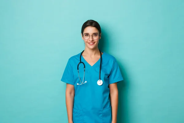 Koronavírus, világjárvány és társadalmi távolságtartás. Kép magabiztos női orvos mosolyog, visel köpeny és szemüveg, pózol a kék háttér — Stock Fotó