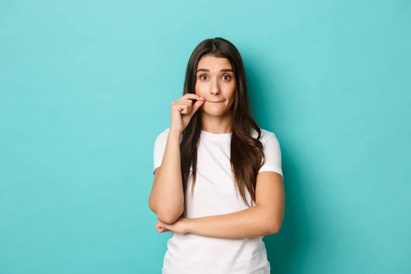 Ritratto di donna bruna carina in t-shirt bianca che fa promesse, labbra sigillate, bocca con cerniera per nascondere il segreto, in piedi su sfondo blu — Foto Stock