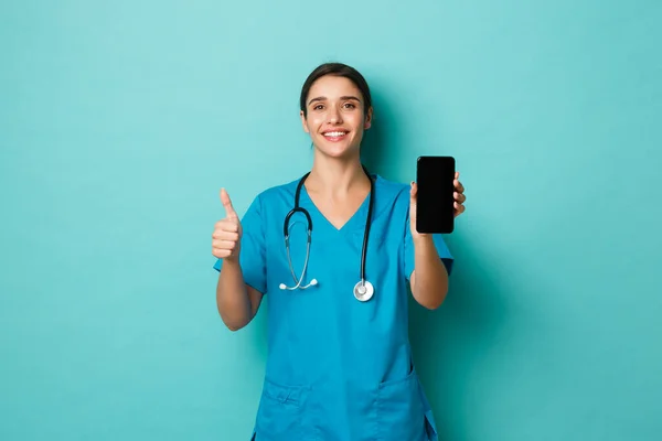 Covid-19, pandemia y concepto de medicina. Imagen de la bella doctora sonriente, vistiendo uniformes, mostrando la pantalla del teléfono móvil y los pulgares hacia arriba, recomiendan la aplicación, fondo azul — Foto de Stock