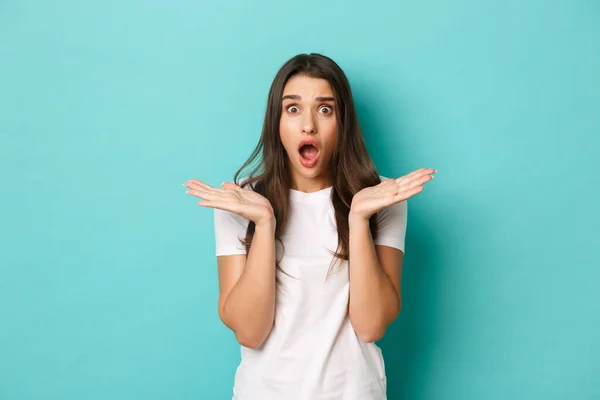 Image of shocked and anxious brunette woman in white t-shirt, spread hands sideways and gasping alarmed, standing over blue background — Stock Photo, Image