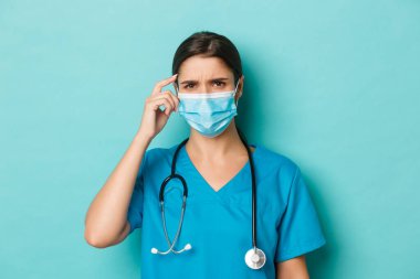 Covid-19 ve karantina kavramı. Tıbbi maskeli, önlüklü, kaşlarını çatmış, şaşkın ve mavi arka planda duran kafası karışmış kadın doktorların yakın çekimi.