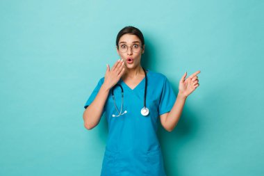 Coronavirus, salgın hastalık ve sosyalleşme konsepti. Şaşırmış ve şaşırmış güzel bir doktorun görüntüsü, önlük ve gözlük takmış, parmağıyla fotokopi alanını işaret ediyor, mavi arka plan.