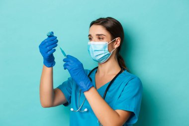 Covid-19 ve karantina kavramı. Tıbbi maskeli, eldivenli ve önlüklü kadın doktor görüntüsü. Şırıngayı koronavirüs aşısıyla dolduruyor. Mavi arka planda duruyor.