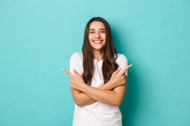 Beyaz tişörtlü neşeli, güzel bayan öğrenci, gülümseyen, mutlu ve parmaklarını yana doğru gösteren, mavi fotokopi alanında iki varyant gösteriyor.