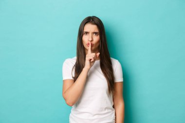 Kızgın esmer kadın resmi, beyaz bir tişört giyiyor, şşşş yapıyor ve yüksek sesle bağırıyor, mavi arka planda duruyor.