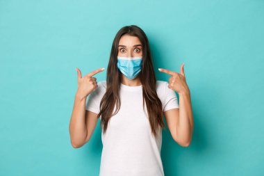 Salgın, covid-19 ve sosyalleşme kavramı. Beyaz tişörtlü harika bir kız, mavi arka planda duran Coronavirus sırasında tıbbi maske takmayı öneriyor.