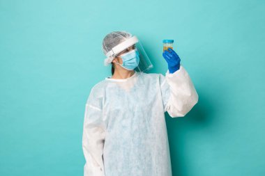 Koronavirüs, sağlık ve karantina kavramı. Analiz etmek için hastanın idrar örneğine bakan, mavi arka planda duran, kişisel koruyucu ekipmanlı kadın doktor görüntüsü.