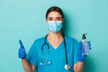Covid-19 ve karantina kavramı. Tıbbi maskeli, eldivenli ve önlüklü güzel kadın doktorlara yakın çekim. Baş parmaklarını gösteren önlükler. Coronavirüs 'ten korunmak için el dezenfektanı kullanma tavsiyesi.