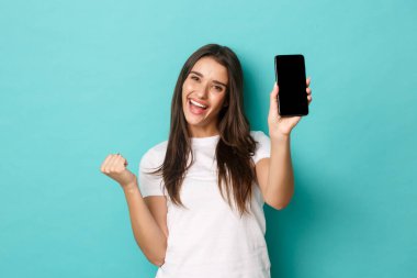 Başarılı ve mutlu bir kadının portresi, akıllı telefon ekranı göstermek ve kazanmak için yumruk hareketi yapmak, uygulamada amaca ulaşmak, mavi arka planda durmak.