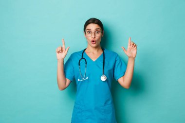 Coronavirus, salgın hastalık ve sosyalleşme konsepti. Önlük ve gözlüklü heyecanlı kadın stajyer resmi, steteskopu boynuna dola, parmaklarını logoya kaldır, mavi arka planda dur.