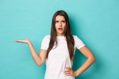 Beyaz tişörtlü, hayal kırıklığına uğramış genç bir kadın bir elini kaldırıyor ve kafası karışmış, mavi arka planda dikiliyor.