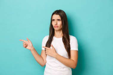 Hüsrana uğramış ve üzgün genç bir kadın görüntüsü, somurtuyor ve somurtuyor, fotokopi alanını işaret ediyor ve sola bakıyor, mavi arka planda duruyor.