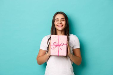 Tatil ve kutlama kavramı. Beyaz tişörtlü, güzel, rüya gibi bir kadın, elinde hediyeyle mutlu bir şekilde gülümsüyor, mavi arka planda dikiliyor.