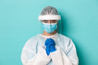 Covid-19, salgın ve karantina kavramı. Kişisel koruyucu ekipmanlarla umut dolu kadın doktorların yakın çekimleri, dua ederken, yalvarırken, yalvarırken, mavi arka planda dururken.