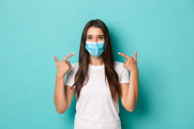 Salgın, covid-19 ve sosyalleşme kavramı. Beyaz tişörtlü güler yüzlü kız Coronavirus sırasında maske takmayı öneriyor. Mavi arka planda duruyor.