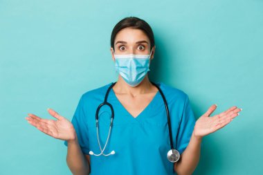 Covid-19 ve karantina kavramı. Tıbbi maskeli, önlüklü, ellerini yana açıp omuz silken mavi arka planda dikilen şaşırmış kadın doktorların yakın çekimi.