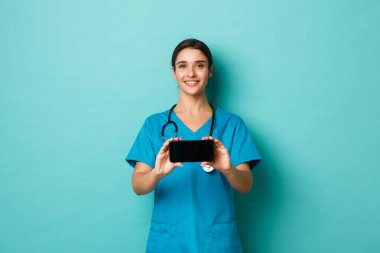 Coronavirus, salgın hastalık ve sosyalleşme konsepti. Önlüklü gülümseyen kadın doktor görüntüsü, akıllı telefonu yatay olarak gösteriyor, mavi arka planda duruyor.