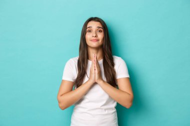 Sevimli ve umut dolu bir kadın imgesi yardım istiyor, gülümsüyor ve el ele tutuşup dua ediyor, mavi arka planda beyaz tişörtle dikiliyor.