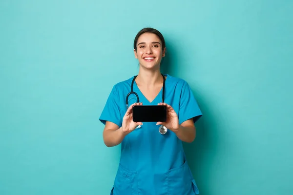 Coronavirus, pandemia y distanciamiento social. Imagen de una doctora sonriente en uniforme, mostrando el teléfono inteligente horizontalmente, de pie sobre fondo azul — Foto de Stock