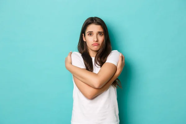 Lonely timid girl in white t-shirt, pouting and looking insecure, hugging herself, seeking comfort, standing over blue background — Stock Photo, Image