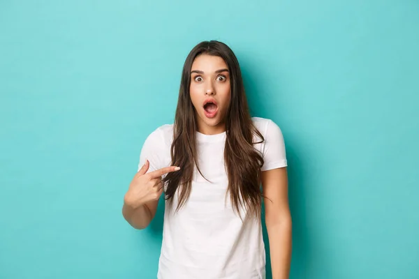 Shocked young woman in white casual shirt, looking startled and pointing at herself, standing surprised over blue background — Stock Photo, Image